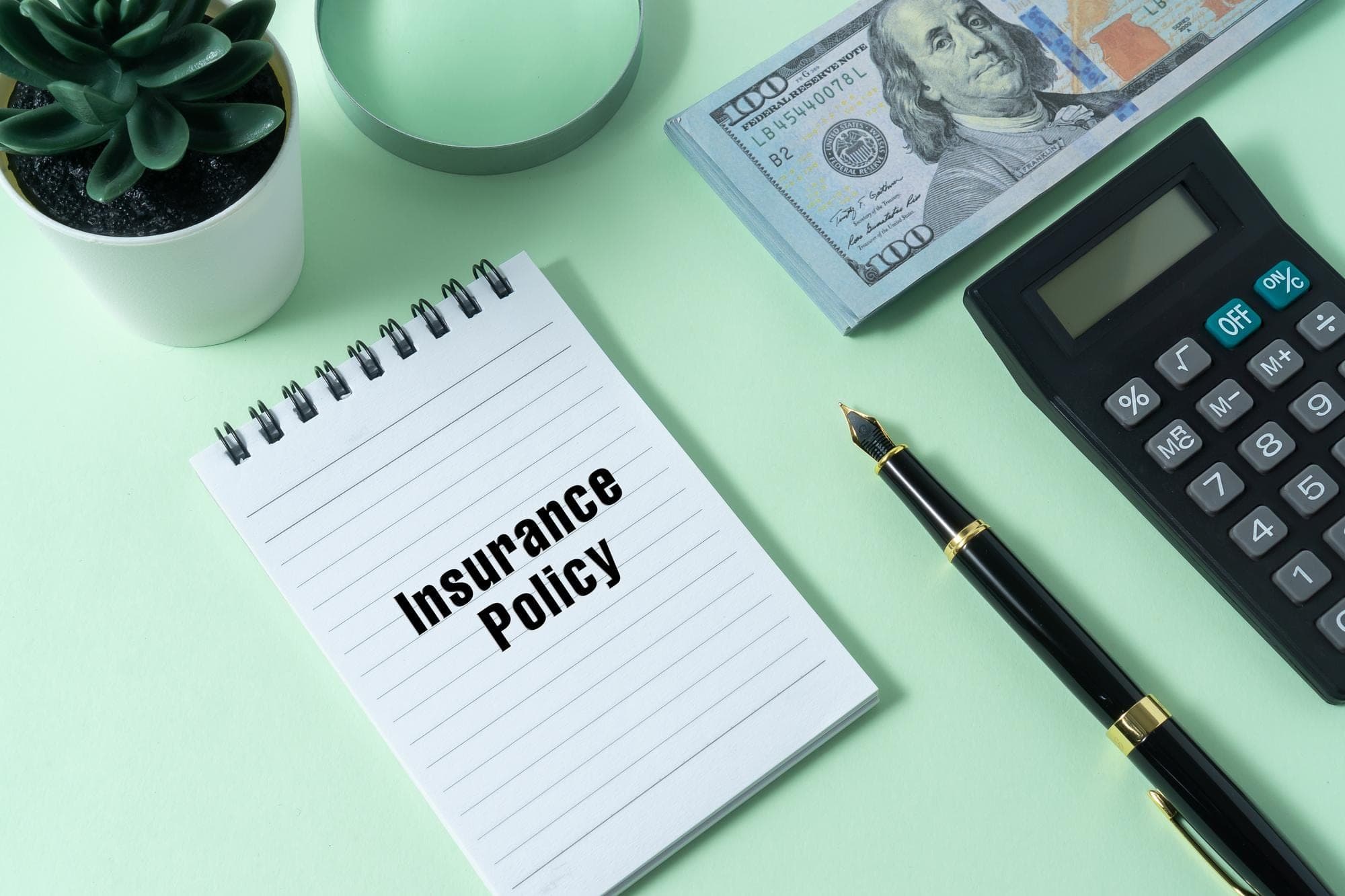 Calculator, pen, plant, money, notebook with 'Insurance Policy' on green background.
